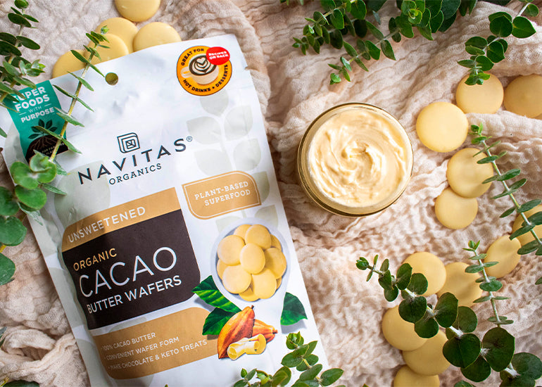 A jar of creamy body butter surrounded by a package and loose Navitas Organics Cacao Butter Wafers and greenery