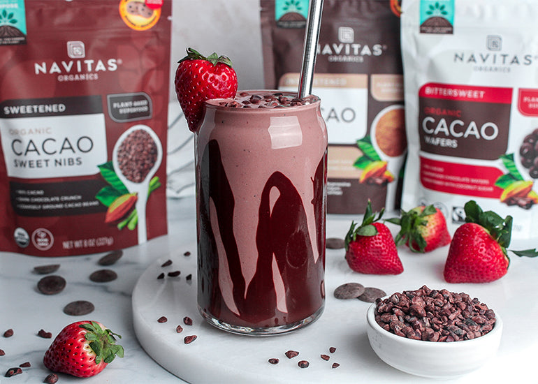 A tall glass lined with melted Navitas Organics Bittersweet Cacao Wafers and filled with a chocolate strawberry smoothie made with Navitas Organics Cacao Powder, topped with Navitas Organics Cacao Sweet Nibs and a fresh strawberry. 