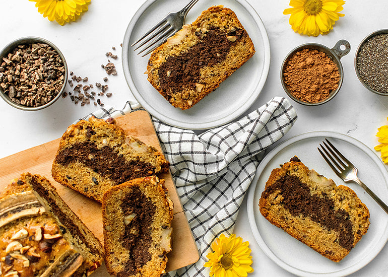 A chocolate hazelnut banana bread with two slices on plates all surrounded by yellow flowers, and measuring cups filled with cacao nibs, cacao powder and chia seeds.