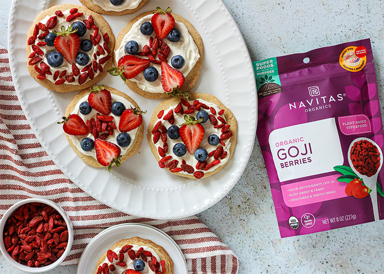A top view of a kitchen counter with a plate of sugar cookies topped with cream cheese, strawberries, blueberries, and goji berries. A bowl of goji berries and a package of Navitas Organics Goji Berries are also visible.