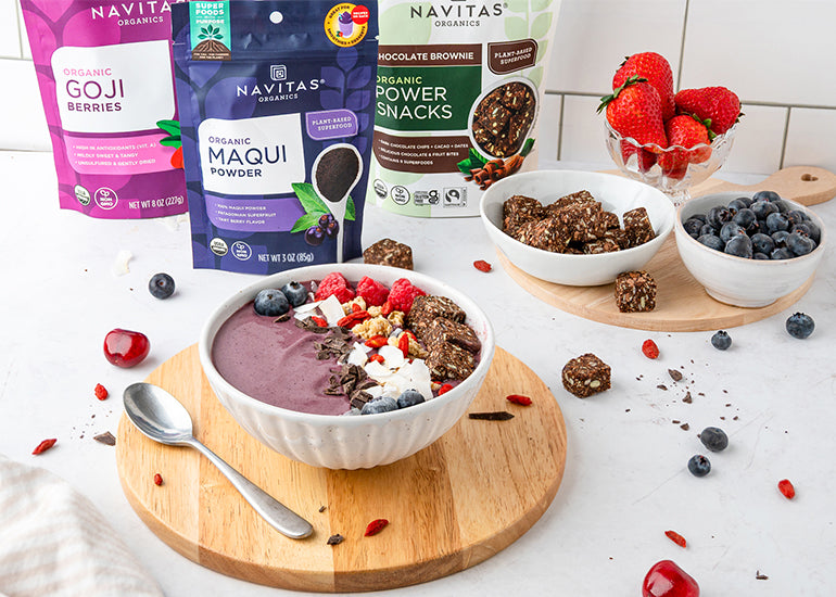 A maqui berry smoothie bowl topped with fruit, nuts and seeds, accompanied by two wooden plates holding fresh berries and chocolate snacks. In the background are bags of Navitas Organics Maqui Powder and Chocolate Brownie Power Snacks. 