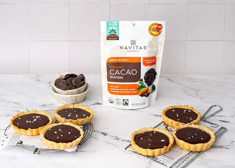 A package of Navitas Organics Semi-Sweet Organic Cacao Wafers stands on a kitchen counter next to a bowl of cacao wafers and a cooling rack with six chocolate tarts.