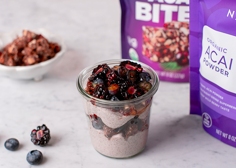 A glass jar filled with layers of yogurt and crumbled snack bites topped with mixed berries, next to packages of Navitas Organics Açaí Powder and Berry Acai BITES.