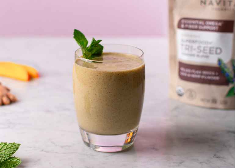 A glass of smoothie garnished with a mint sprig sits on a marble surface. In the background, there's a blurred bag of Navitas Organics Superfood+ Tri-Seed Powder Blend. Mango slices and ginger are visible on the left.