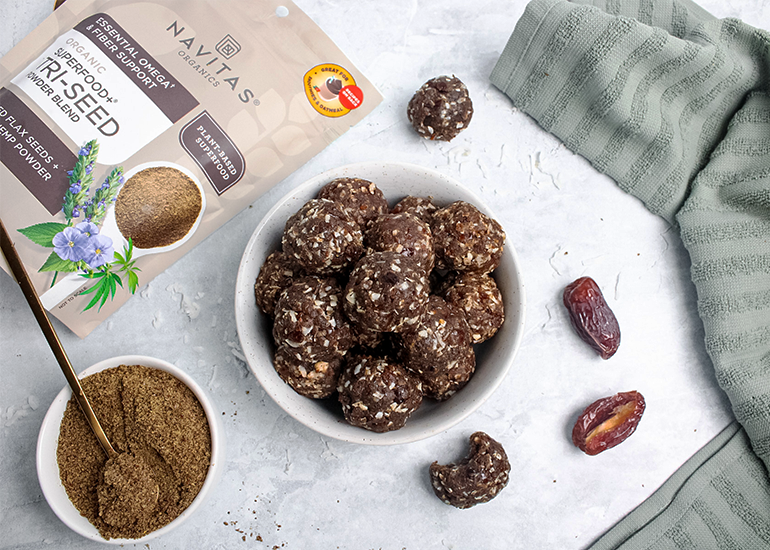 A bowl of date-based energy balls sits on a gray surface surrounded by dates, a bag of Navitas Organics Superfood+ Tri-Seed Powder Blend and a bowl of the powder with a gold spoon.