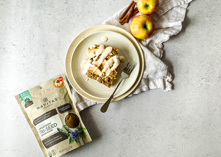 A plate of apple spice oat bars drizzled with icing, accompanied by a fork. Nearby are apples, cinnamon sticks, and a package of Navitas Organics Superfood+ Tri-Seed Powder Blend. A napkin is partially visible under the items.