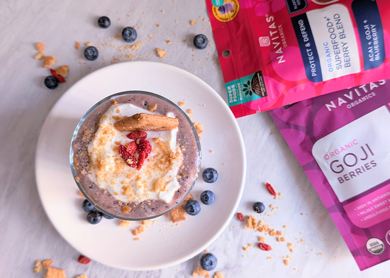A smoothie topped with goji berries, blueberries, yogurt and crumbled graham crackers next to bags of Navitas Organics Goji Berries and Superfood+ Berry Blend, surrounded by scattered blueberries and graham cracker crumbs on a marble surface.