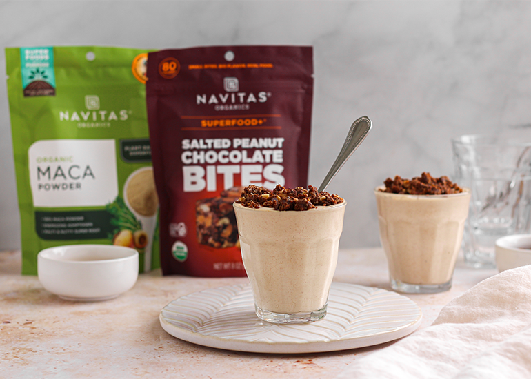 Two glasses filled with a creamy smoothie, topped with crumbled snacks and spoons inside. Behind them, packages of Navitas Organics Maca Powder and Salted Peanut Chocolate BITES are visible. A few empty bowls and glasses are on the side.