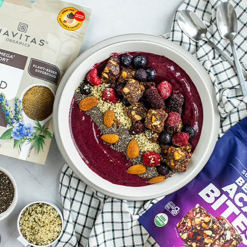 An acai smoothie bowl topped with a variety of nuts, seeds, and berries is surrounded by bags of Navitas Organics Superfood Tri-Seed Powder Blend and Berry Acai Bites. Additional small bowls contain seeds, alongside two checkered napkins on the surface.