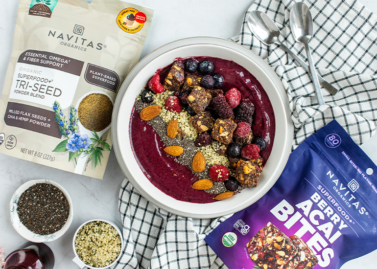 An acai smoothie bowl topped with a variety of nuts, seeds, and berries is surrounded by bags of Navitas Organics Superfood Tri-Seed Powder Blend and Berry Acai Bites. Additional small bowls contain seeds, alongside two checkered napkins on the surface.