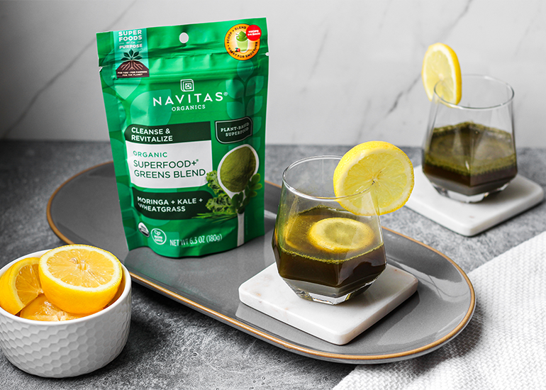 A package of Navitas Organics Superfood+ Greens Blend next to a glass of dark green juice with a slice of lemon, on a tray with fresh lemon halves nearby.