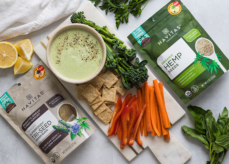 Packages of Navitas Organics Hemp Seeds and Superfood Tri-Seed Powder Blend are placed on either side of a bowl of green dip, carrot and bell pepper sticks, broccoli, crackers, lemon wedges, and fresh basil leaves.
