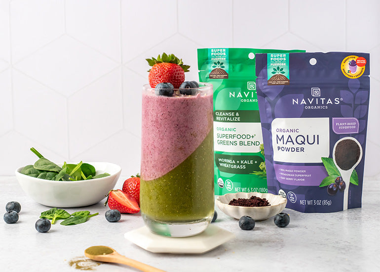 A layered berry and greens smoothie made with Navitas Organics Superfood+ Greens Blend and Maqui Powder in a tall glass topped with a fresh strawberry and blueberries surrounded by blueberries, fresh mint leaves and bags of Navitas Organics superfoods.