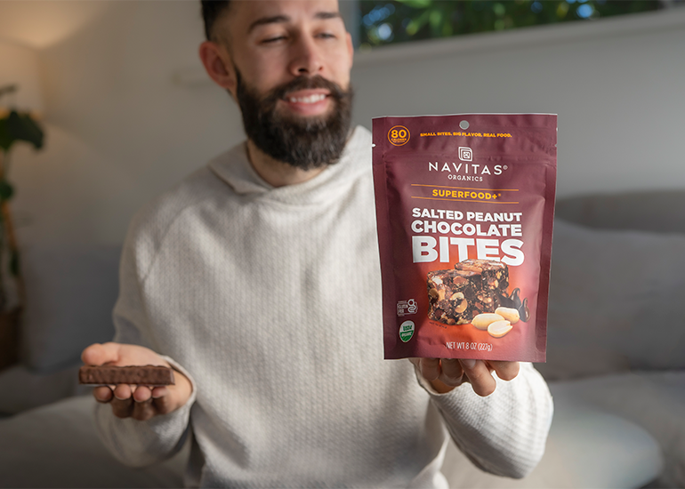 Person holding a package of Navitas Organics Superfood+ Salted Peanut Chocolate BITES in one hand and a chocolate-covered candy bar in the other, while looking at the BITES and smiling in a bright, cozy indoor setting.